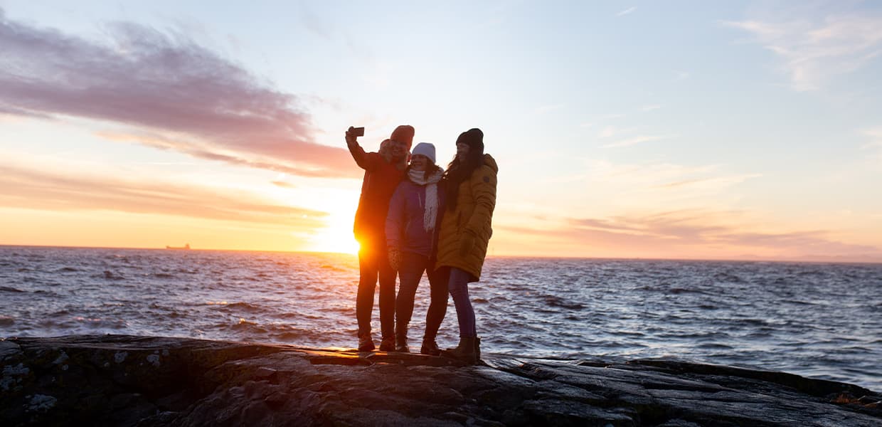 Tre personer står på en klippe og tar en selfie med solnedgang over havet i bakgrunnen.