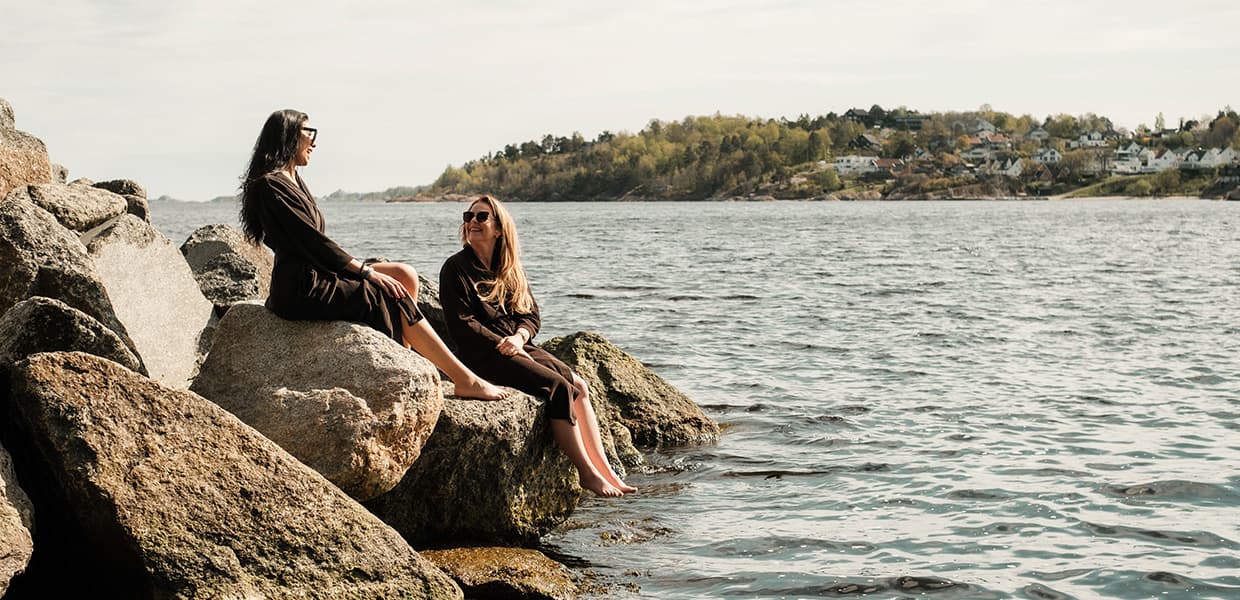 to jenter sitter på steiner med beina som dingler ned i sjøen