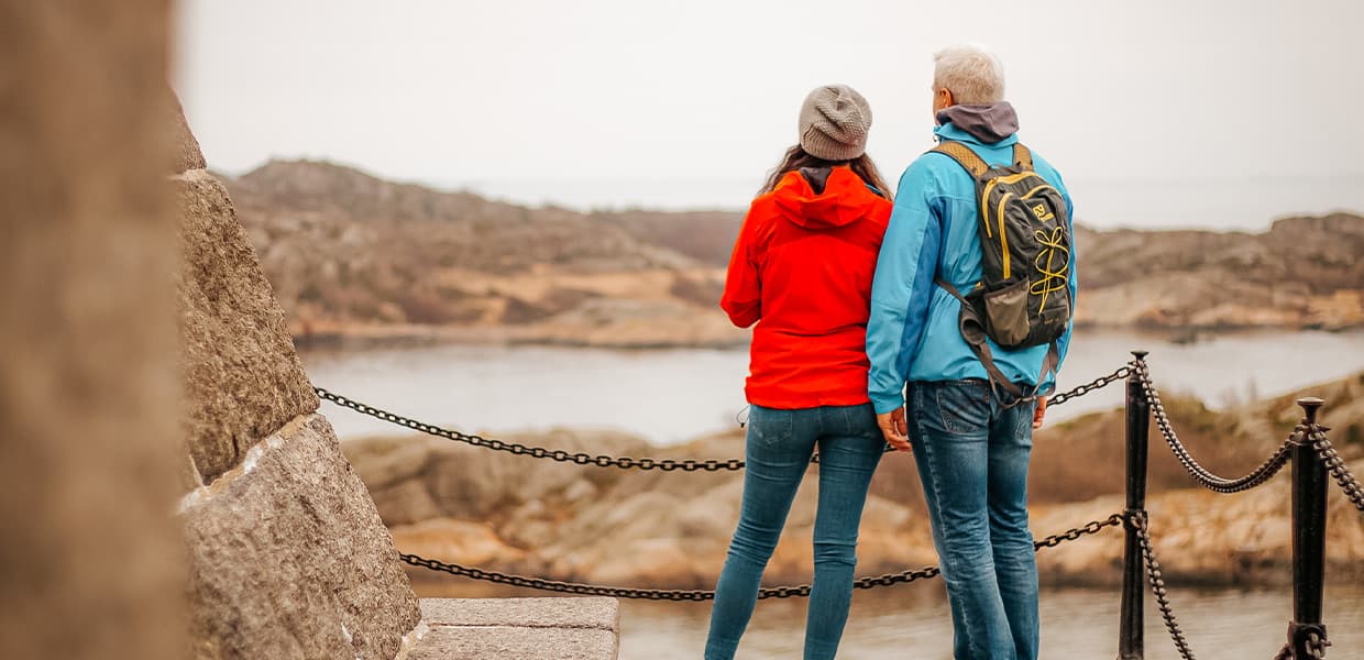 Ei i rød jakke og en i blå jakke med sekk står ved siden av hverandre og ser utover sjøen, med et sjøfartsmonument på deres venstre side