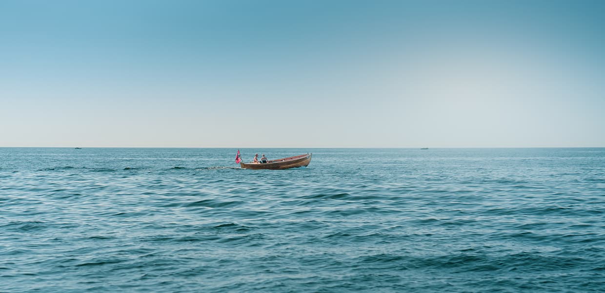 En liten trebåt med norsk flagg seiler på blått hav under klar himmel.