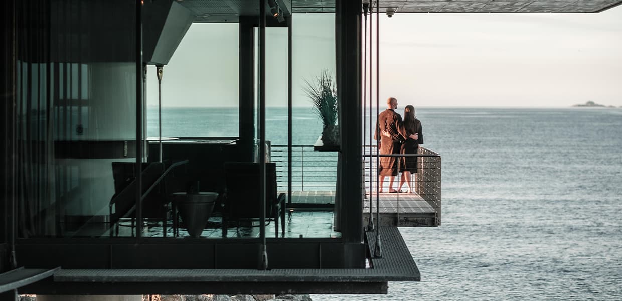 To personer på balkong ved spa med utsikt over havet. Moderne glassfasade med rolig blått hav i bakgrunnen.