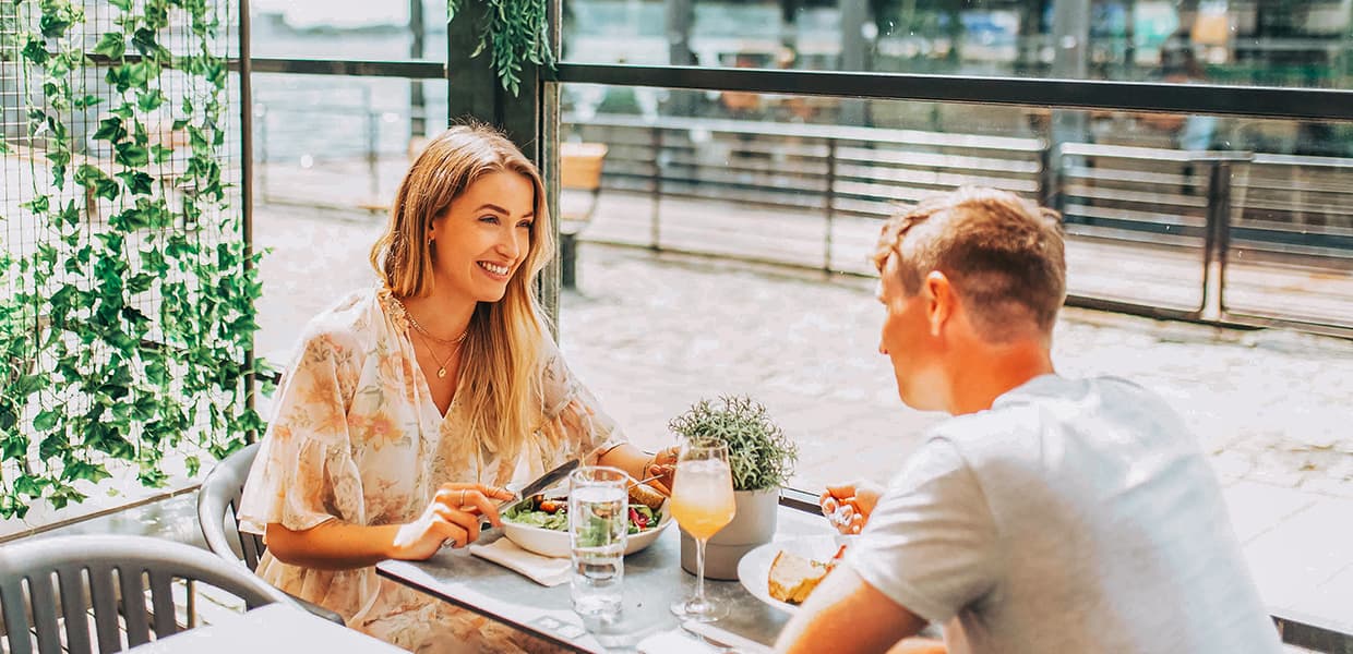 : Et ungt par nyter et lunsjmåltid på en lys og luftig uteservering med store vinduer mot vannet. Den avslappede atmosfæren forsterkes av grønne planter og naturlig lys, mens ferske salater, brød og friske drinker står på bordet. Stemningen er sommerlig og uformell, perfekt for en hyggelig matopplevelse ved bryggekanten.