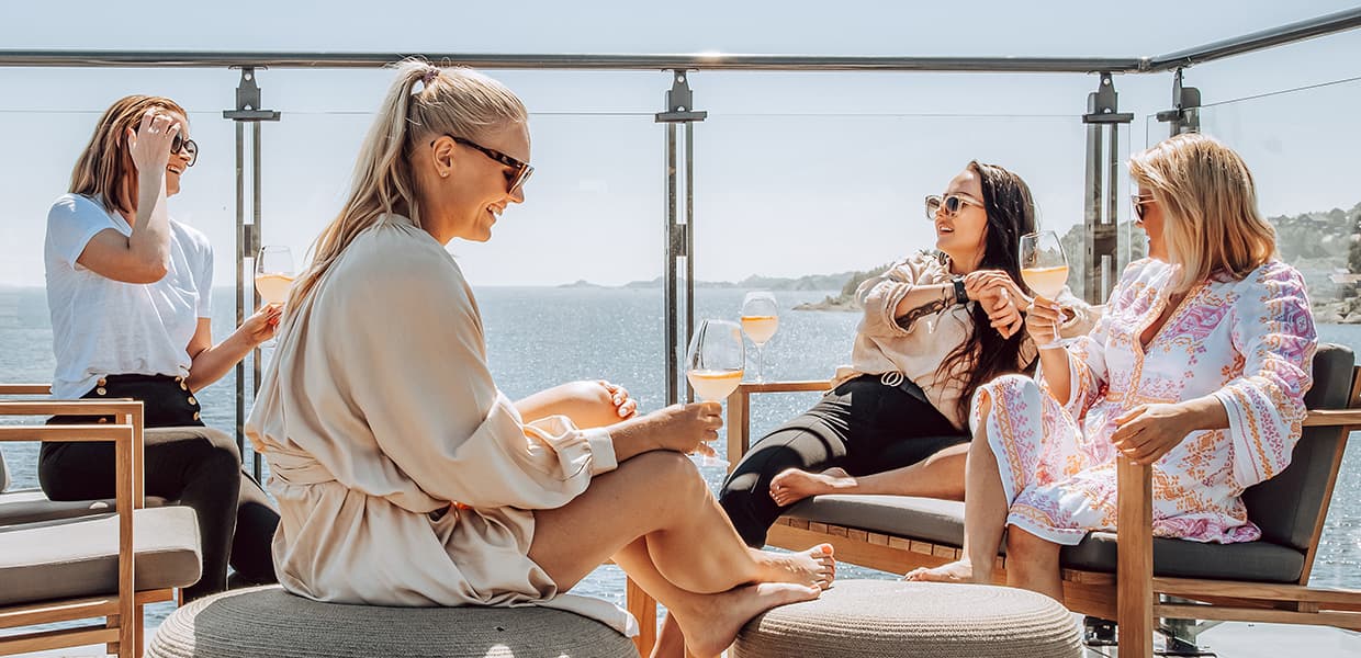 fire damer med vært sitt vinglass med lys drikke sitter og prater på utemøbler på takterassen til Farris Bad. et gjennomsiktig gjerde skiller dem og den glitrende sjøen bak.