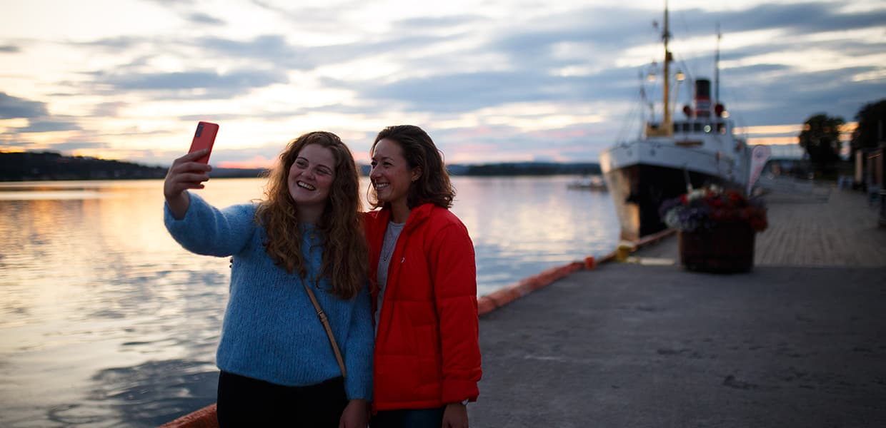 To personer tar en selfie på en brygge med et skip i bakgrunnen i solnedgangen.