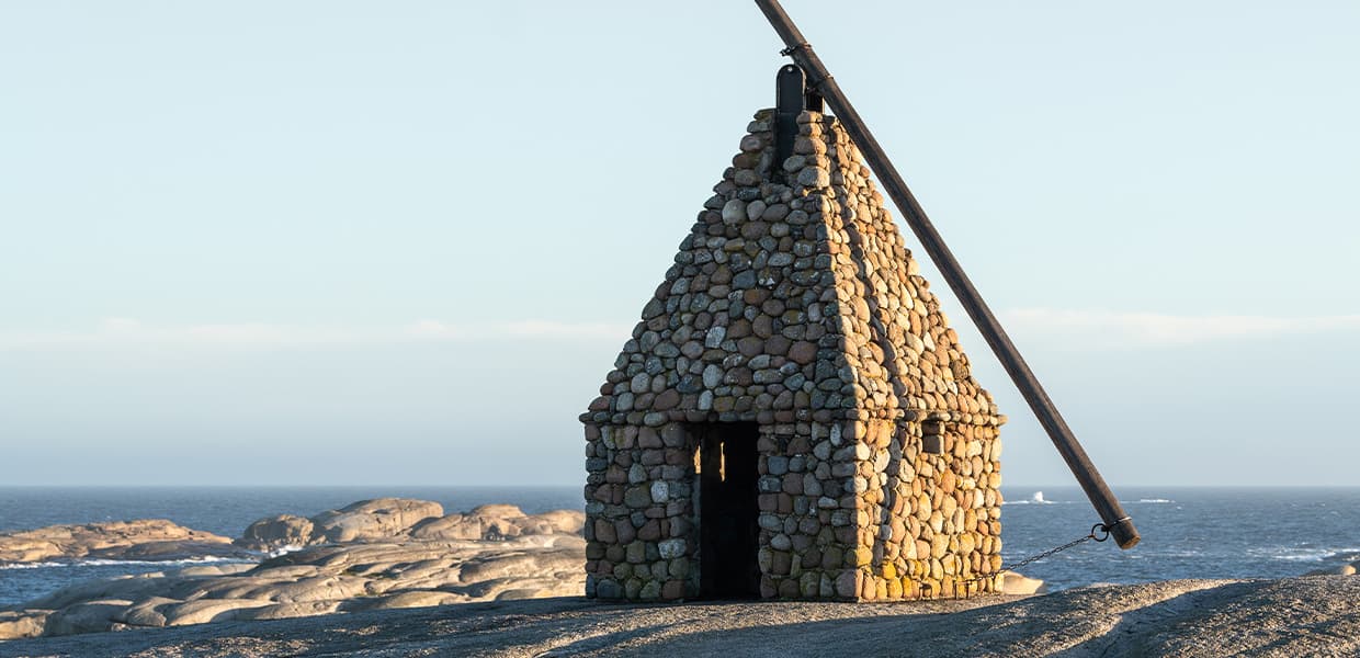 Et fyr bygget av små steiner plassert ved sjøen på Verdens Ende. Under himmelen stikker trekonstruksjonens topp opp mot havet, omgitt av svaberg, med sjøen i bakgrunnen.