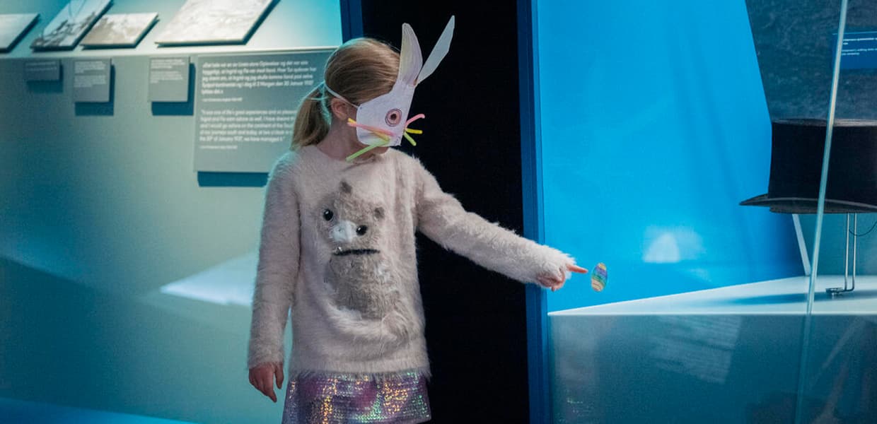 Et barn i rosa, pusete genser med påskeharemaske besøker en mørk utstilling opplyst med blått lys og peker å et påskegg som er klistret på et utstillingsmonter.