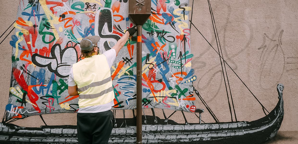 Graffiti-vegg med vikingskip: Person i refleksvest maler fargerikt graffiti på vegg, med en tradisjonell vikingbåt i forgrunnen.