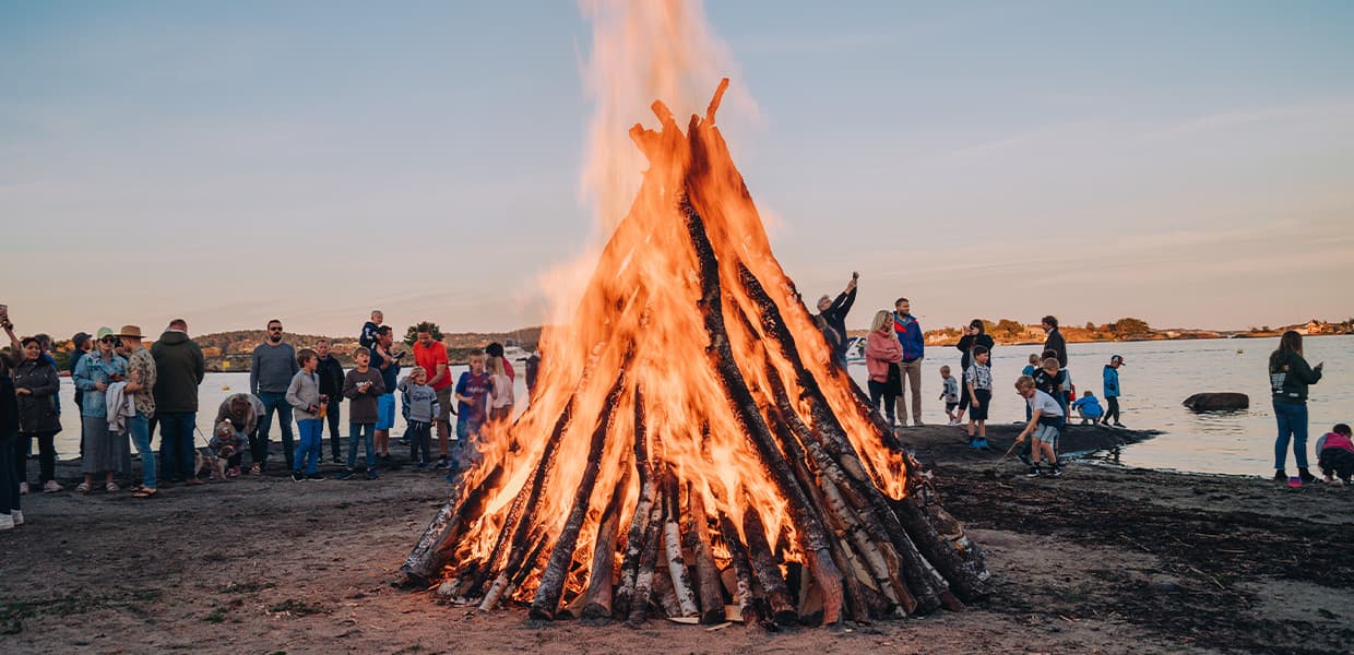 Sankthansbål ved kysten i skumring. Publikum står samlet og ser på de høye flammene. Rosa-oransje himmel og mørke silhuetter av trær i bakgrunnen.