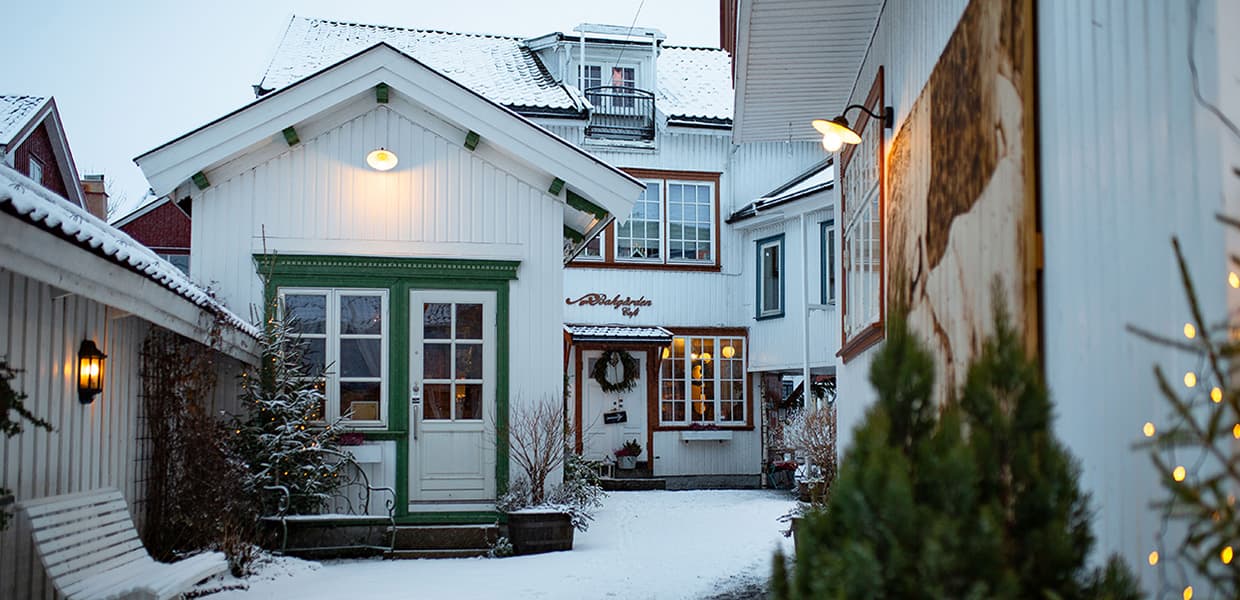 En vinterscene fra en koselig bakgård med tradisjonelle hvite trehus. Inngangene er prydet med grønne dørrammer og juledekorasjoner som kranser. Snøen dekker bakken og takene, mens varme lykter og vinduer lyser opp den dempede vinterkveldstemningen. Et juletre eller en gran står i forgrunnen, og små julelys glitrer langs husveggene. Arkitekturen har typisk nordisk stil med panel og sprossevinduer.