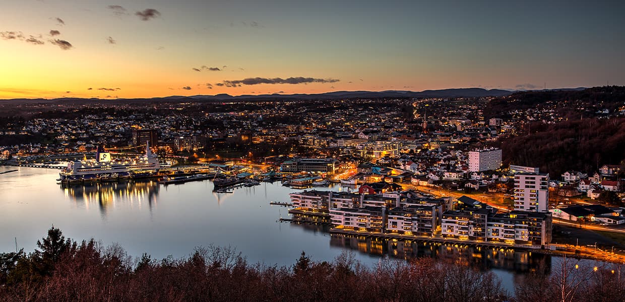 Panoramautsikt over Sandefjord by i skumringen. Byens lys glitrer langs havnen og i bebyggelsen som strekker seg oppover åssidene. Cruiseskip ligger til kai i havnen, og fjorden ligger stille med refleksjoner av byens lys.