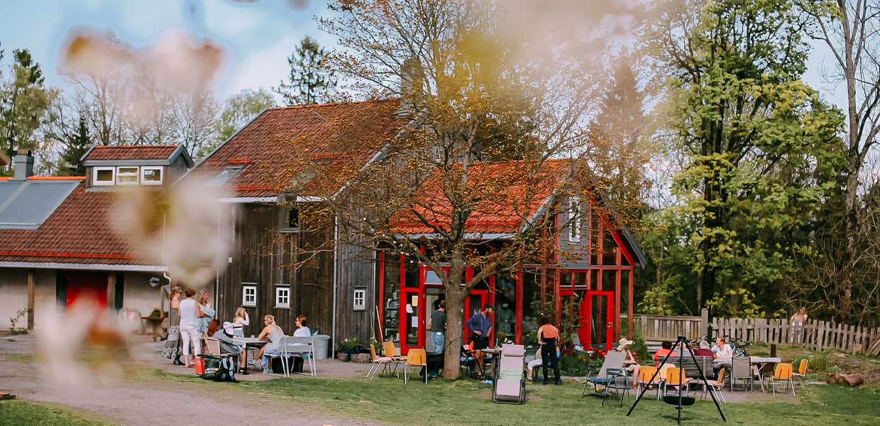 Et tradisjonelt gårdshus med rødt tak og tremmer, omgitt av grønne trær. Folk sitter og prater ved utendørsmøbler.