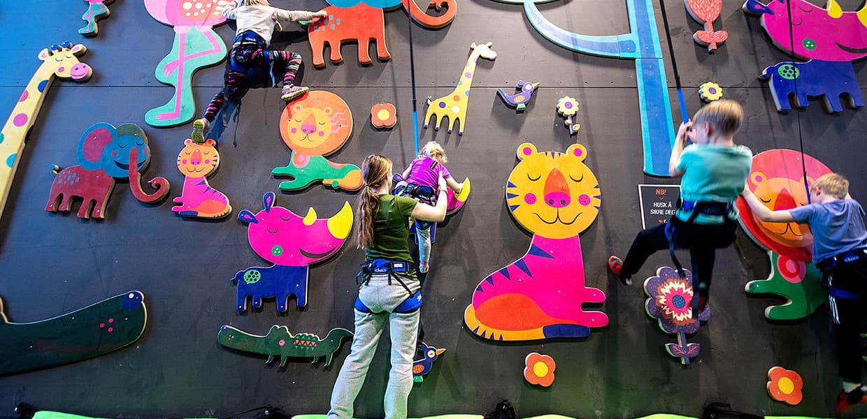 Barn klatrer på en innendørs vegg dekorert med fargerike dyrefigurer som elefant og sjiraff, iført klatreseler.