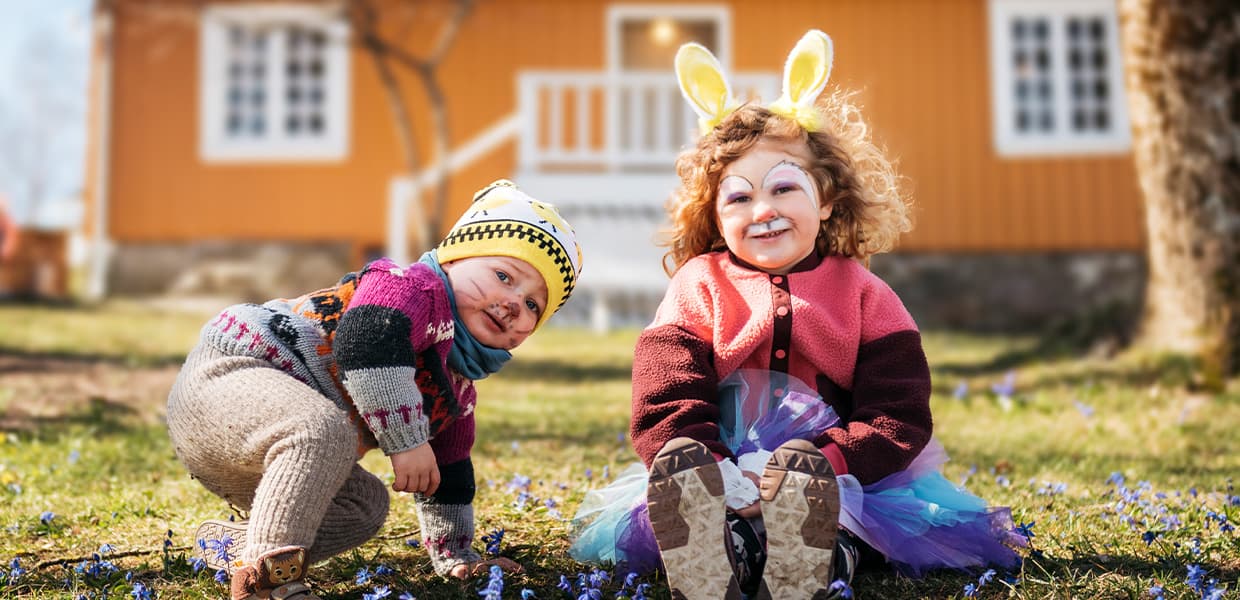 To små barn leker i hagen foran et gult hus. En av barna har påskeharer-ører, og de er omgitt av vakre vårblomster.