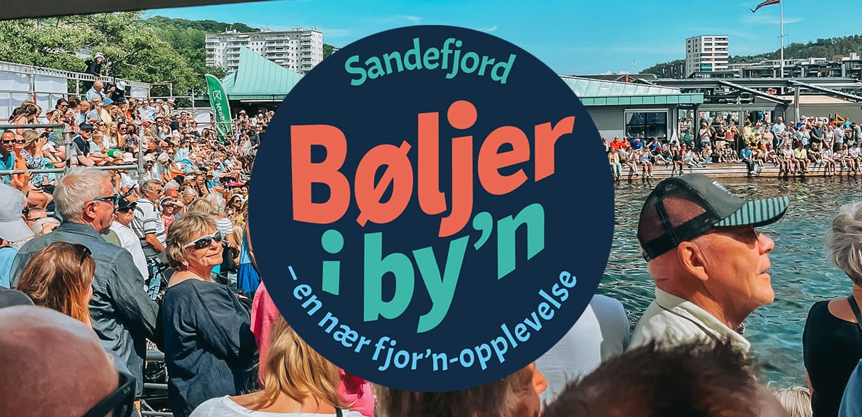 Et stort folkemengde samles ved vannkanten under arrangementet "Bøljer i by'n" i Sandefjord, hvor en svær plakat med arrangementets navn svever i forgrunnen, og publikum ser ut til å nyte den livlige stemningen.