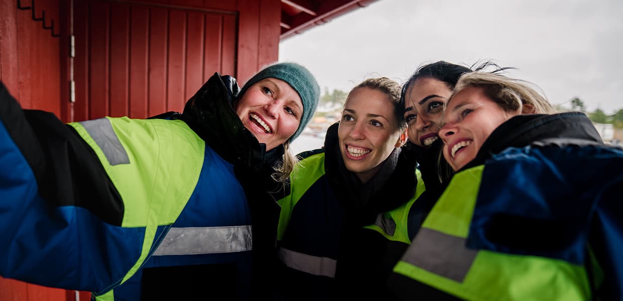 Fire personer i fargerike redningsvester tar en selfie foran en rød sjøbod. De smiler bredt mens de står tett sammen i det som ser ut til å være typisk norsk skjærgårdsmiljø