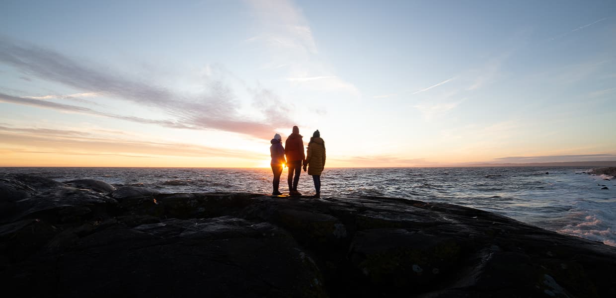 Tre personer står i silhuett på svaberget ved Mølen med havet i bakgrunnen. Solen går ned og skaper en gyllen horisont med pastellfarger på himmelen