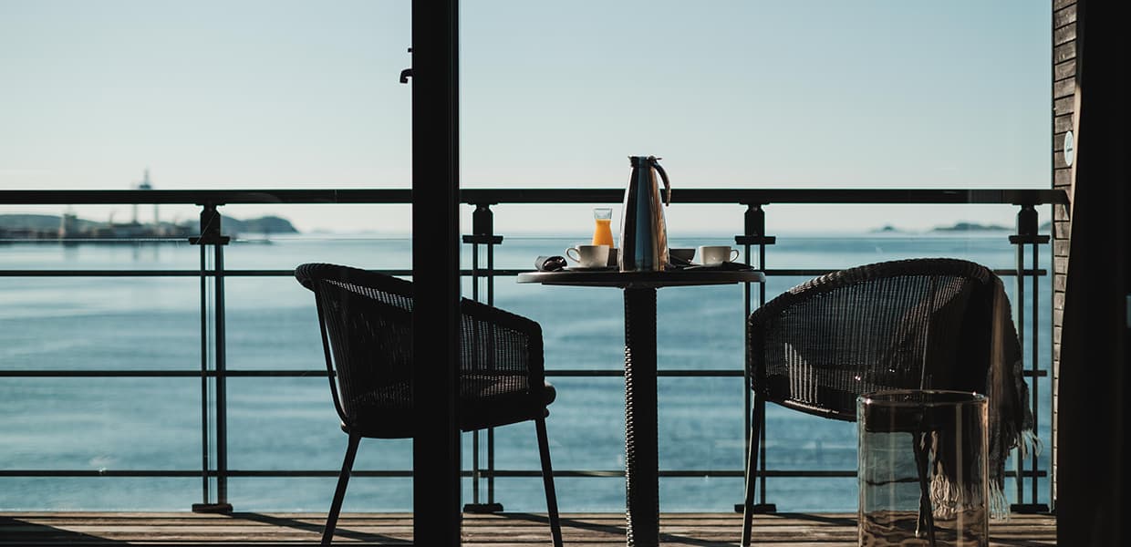 To stoler og et bord dekket med appelsinjuice, termos og kaffekopper står på en solfylt terrasse med sjøutsikt.