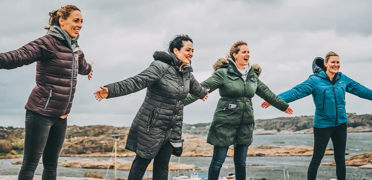 Fire kvinner står på rekke med utstrakte armer ved kysten, med klipper og hav i bakgrunnen.